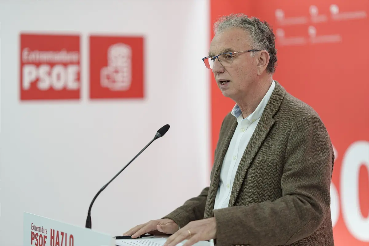 Hombre mayor con gafas y chaqueta marrón hablando en un atril con micrófono en un acto del PSOE de Extremadura con fondo rojo y blanco