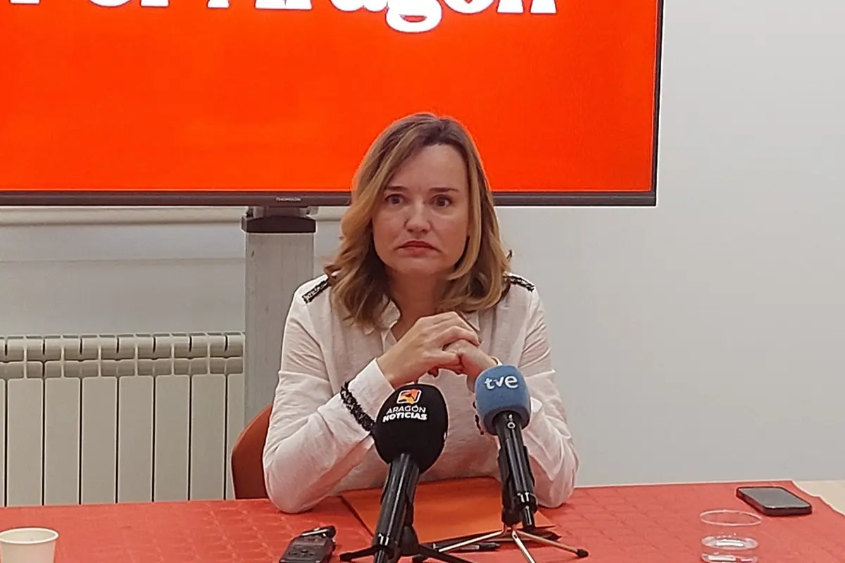 Mujer sentada en una rueda de prensa frente a varios micrófonos con un fondo rojo con texto incompleto Mujer sentada en una rueda de prensa frente a varios micrófonos con un fondo rojo con texto incompleto