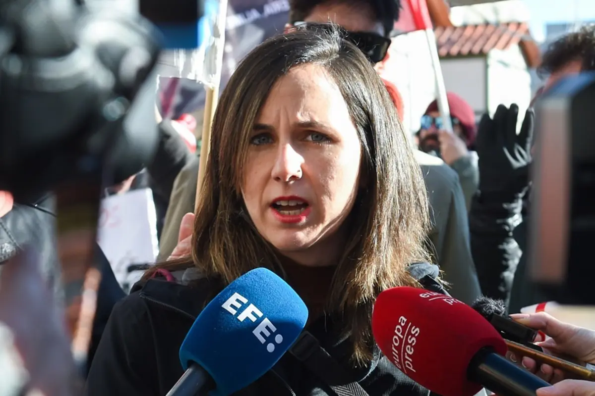 Mujer joven hablando ante varios micrófonos de prensa en una rueda de prensa al aire libre rodeada de personas