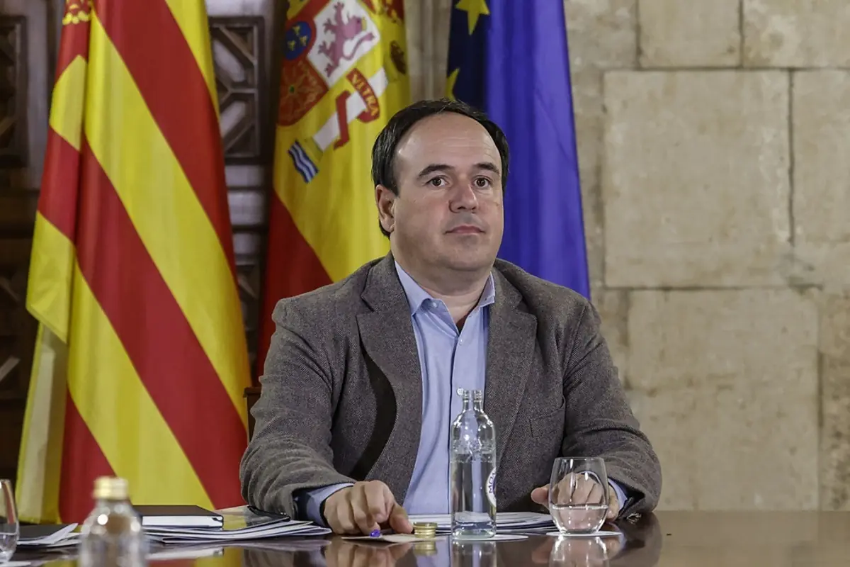 Hombre de mediana edad sentado en una mesa de reuniones con banderas de España la Unión Europea y una comunidad autónoma al fondo