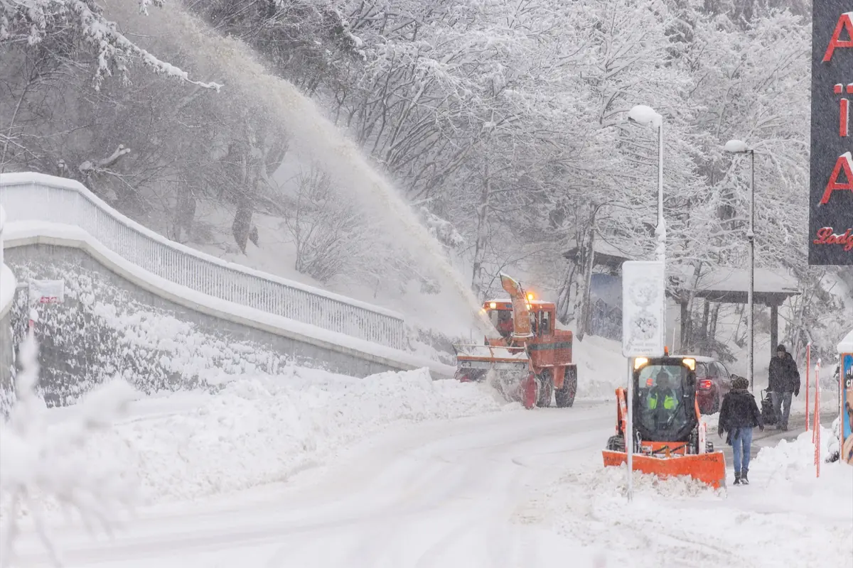Máquinas quitanieves despejan una carretera cubierta de nieve mientras varias personas caminan junto a los montones de nieve y árboles nevados