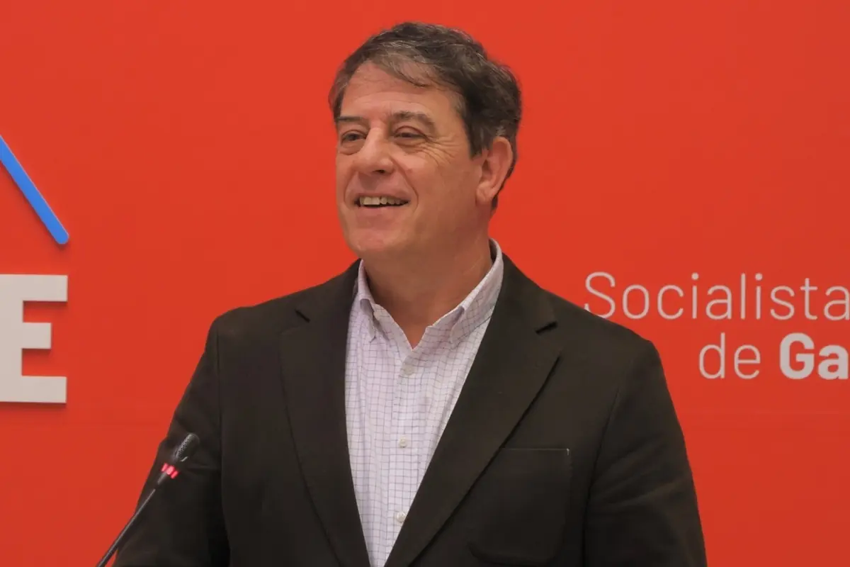 Hombre de mediana edad sonriendo y hablando ante un micrófono con fondo rojo y logotipo de un partido político Hombre de mediana edad sonriendo y hablando ante un micrófono con fondo rojo y logotipo de un partido político
