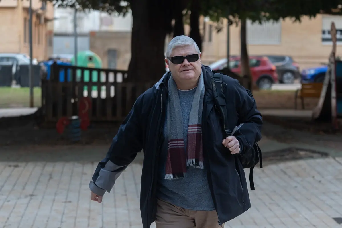 Hombre mayor con gafas de sol caminando por un parque urbano con chaqueta oscura bufanda y mochila