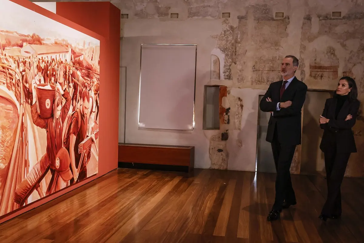 Dos personas elegantemente vestidas observan un gran cuadro de tonos rojizos en una sala de exposiciones con suelo de madera y paredes antiguas