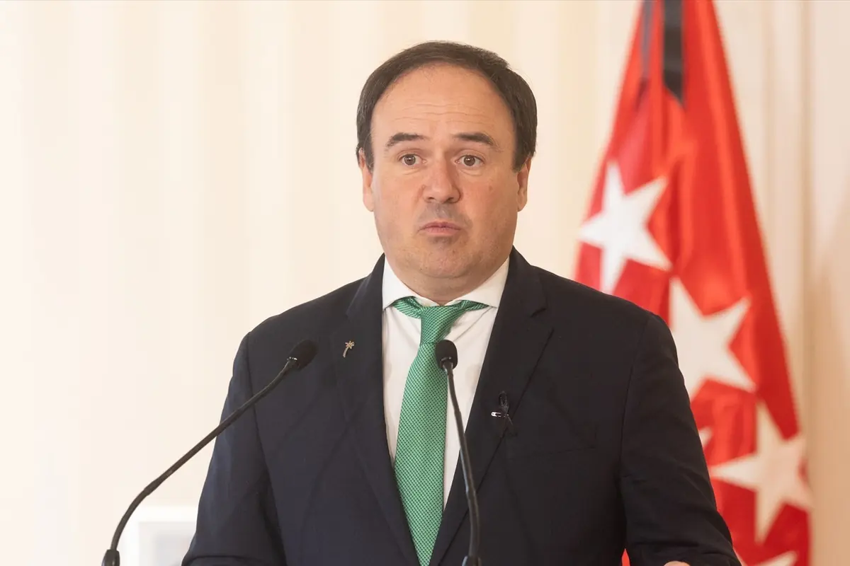 Hombre con traje oscuro y corbata verde hablando frente a dos micrófonos con una bandera roja con estrellas blancas de fondo