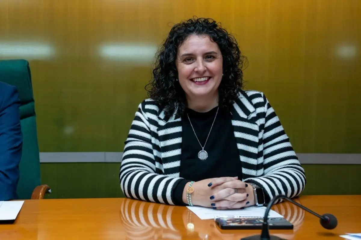 Mujer de pelo rizado y chaqueta de rayas blancas y negras sonriendo sentada en una mesa de reuniones con documentos y un micrófono delante
