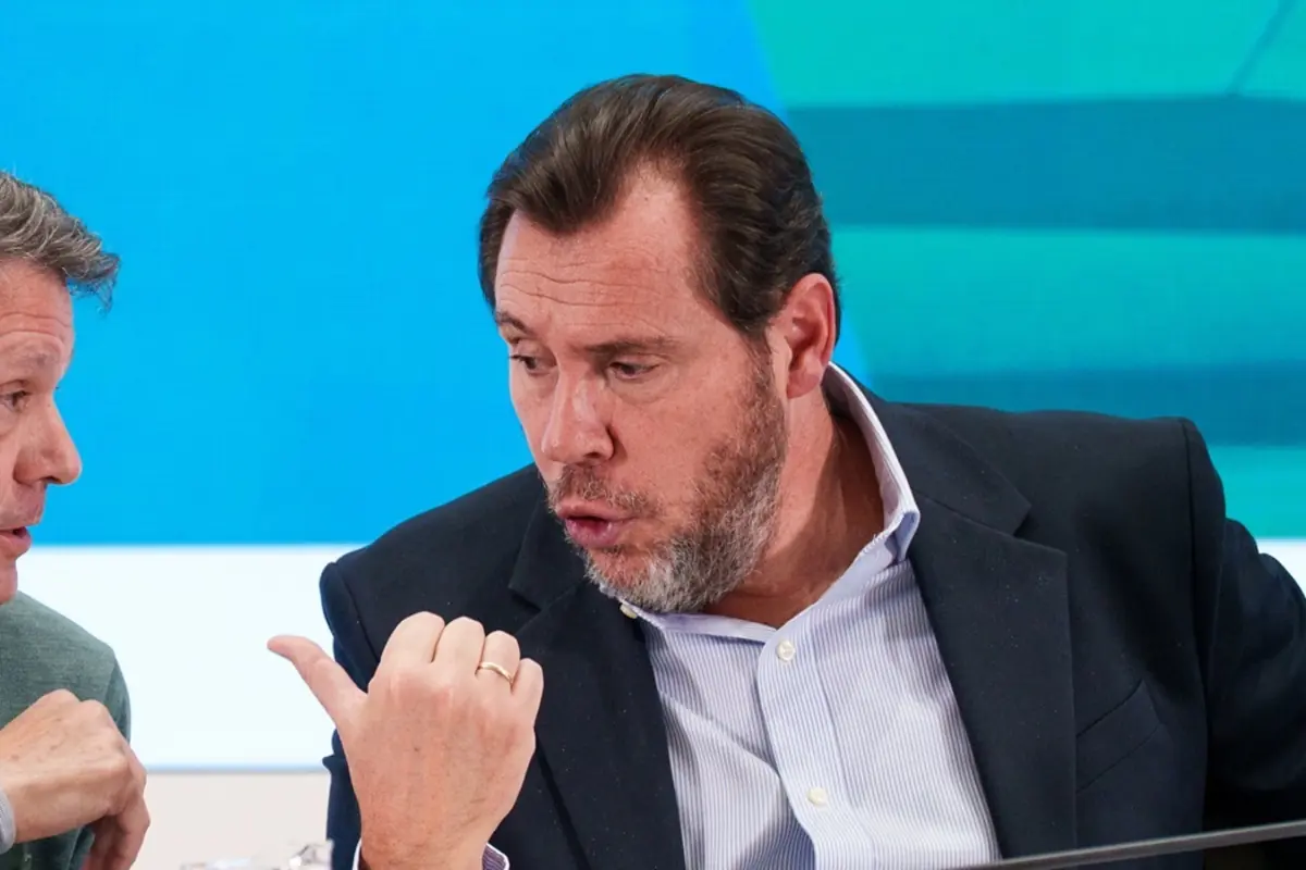 Hombre de mediana edad con barba y traje oscuro gesticulando con la mano mientras conversa con otra persona en un entorno de estudio con fondo azul