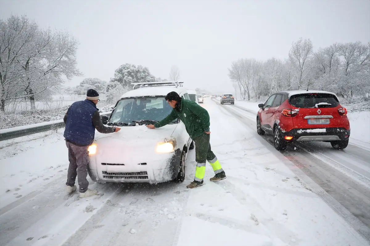 Dos hombres retiran nieve del capó de una furgoneta detenida en una carretera nevada mientras otros coches avanzan con precaución