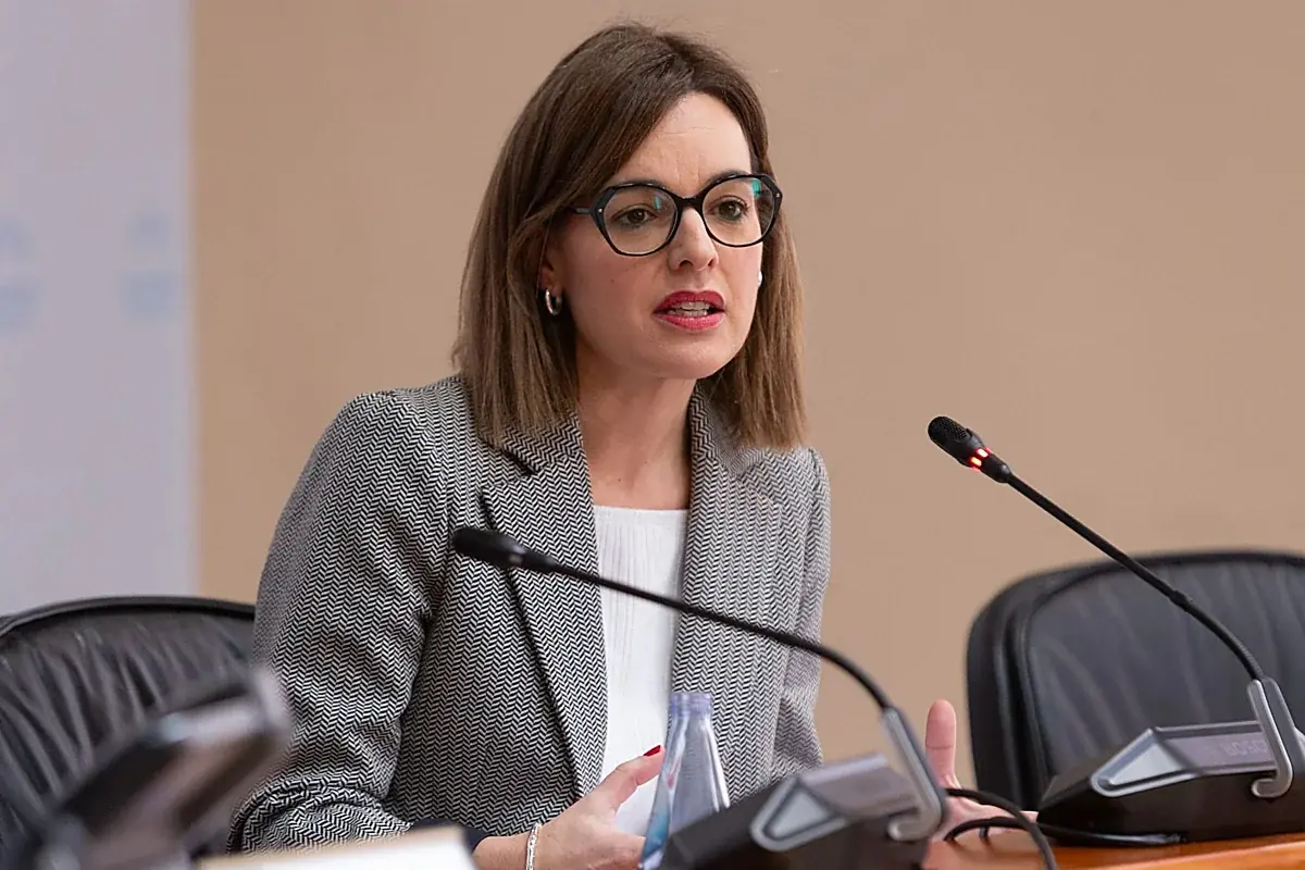 Mujer con gafas y traje gris hablando en una conferencia frente a varios micrófonos