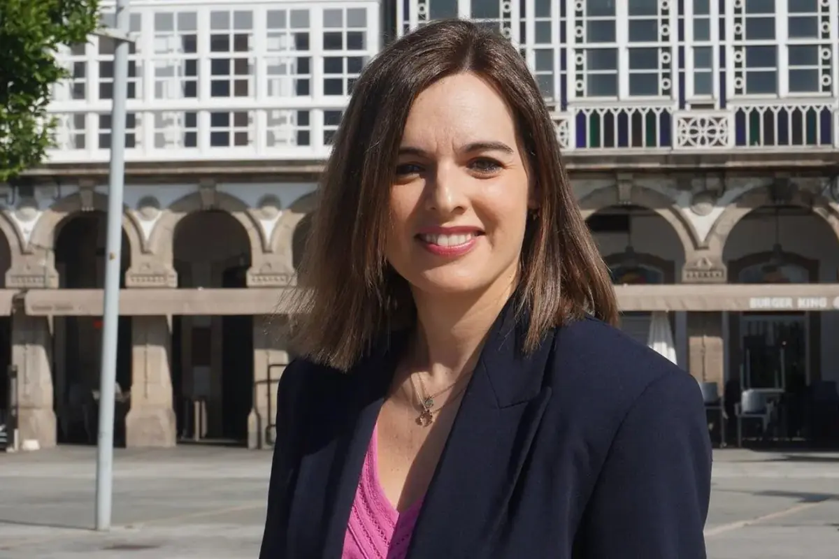 Mujer joven con media melena castaña sonriendo a cámara vestida con blazer azul marino y blusa rosa en una plaza urbana con edificios acristalados al fondo