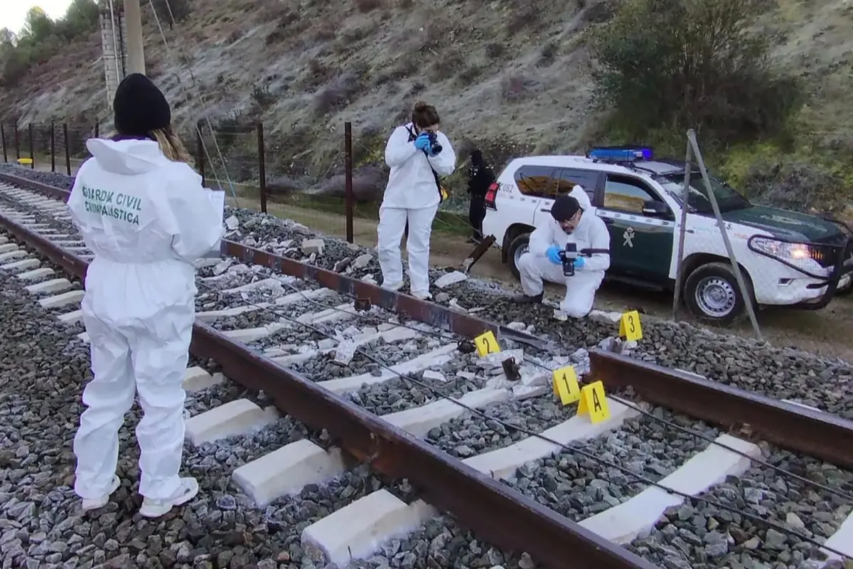 Agentes de la Guardia Civil con trajes forenses inspeccionan unas vías de tren marcadas con señales numeradas junto a un coche patrulla