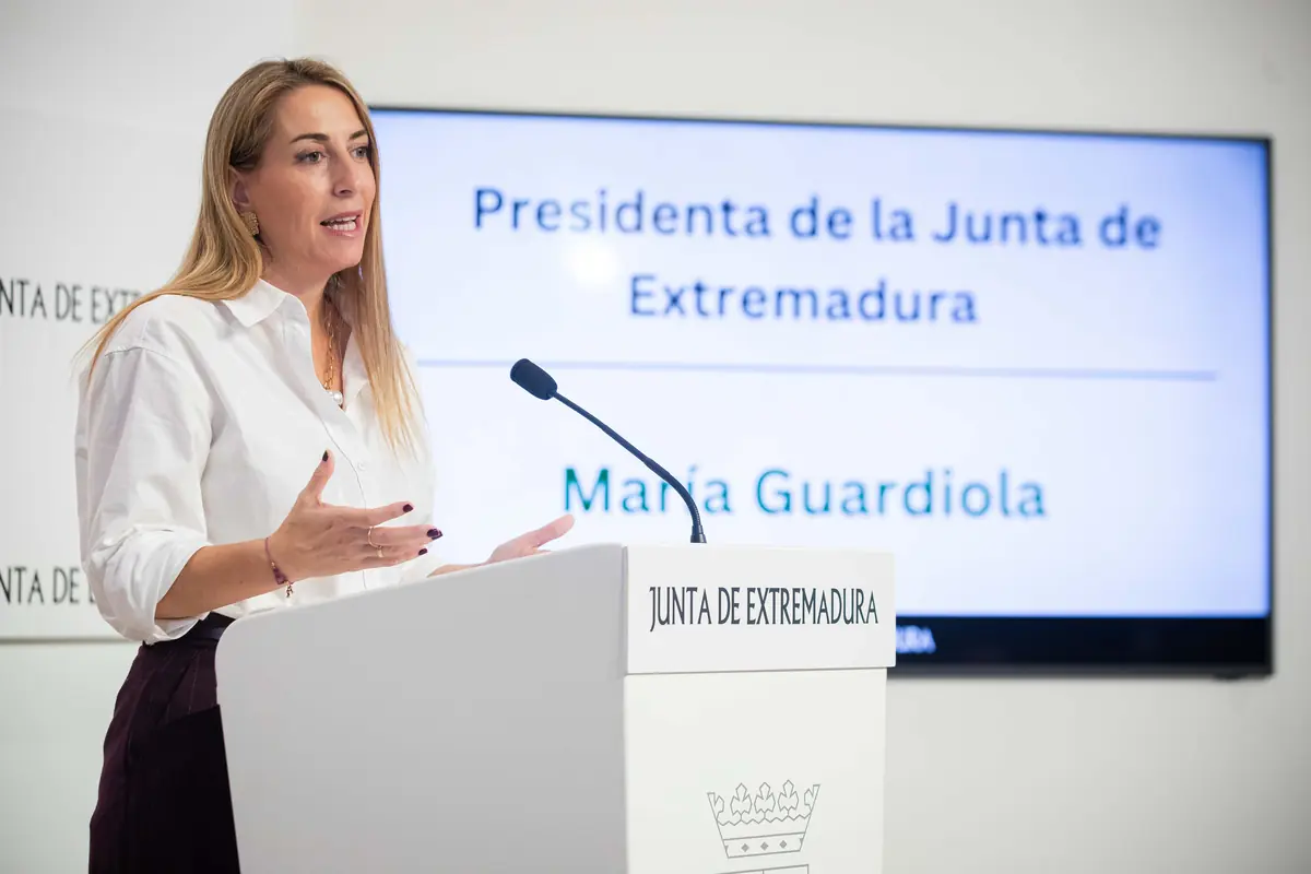 Mujer hablando en un atril de la Junta de Extremadura con una pantalla detrás que muestra el cargo de presidenta de la Junta