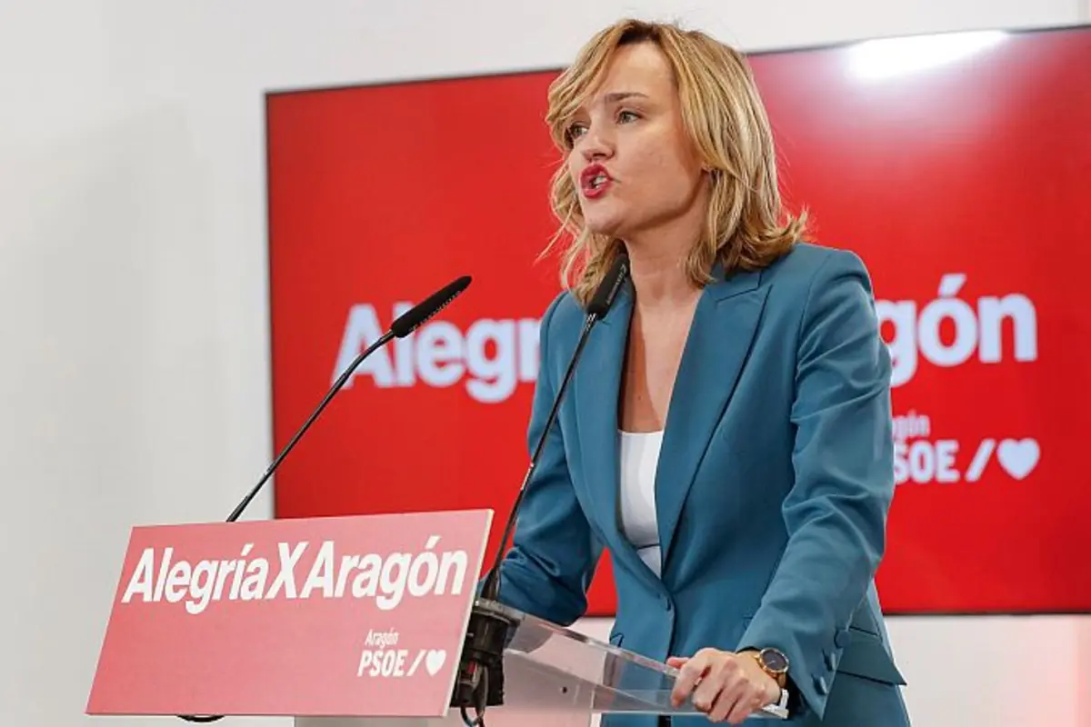 Mujer con traje azul hablando en un atril de campaña con el lema Alegría X Aragón del partido PSOE sobre un fondo rojo