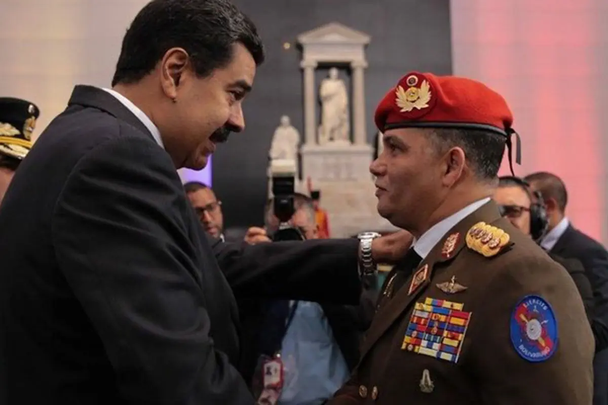 Hombre con traje oscuro estrecha la mano y felicita a un militar con boina roja y uniforme lleno de medallas en un acto oficial con monumentos de fondo