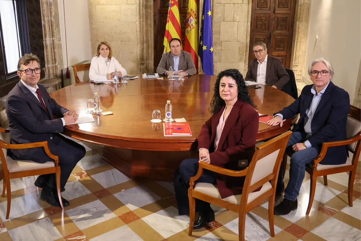 Seis personas sentadas alrededor de una mesa redonda de madera en una sala institucional con banderas oficiales al fondo durante una reunión formal