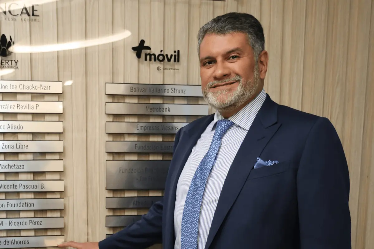Hombre de traje azul sonriendo y señalando una placa con nombres de empresas en una pared de reconocimiento empresarial