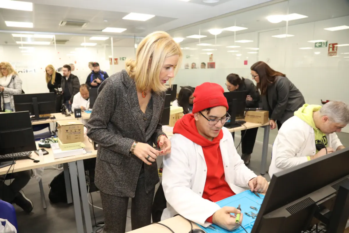 Mujer supervisa a un joven con bata blanca y gorro rojo mientras trabaja con herramientas electrónicas frente a un ordenador en un aula llena de personas y equipos informáticos