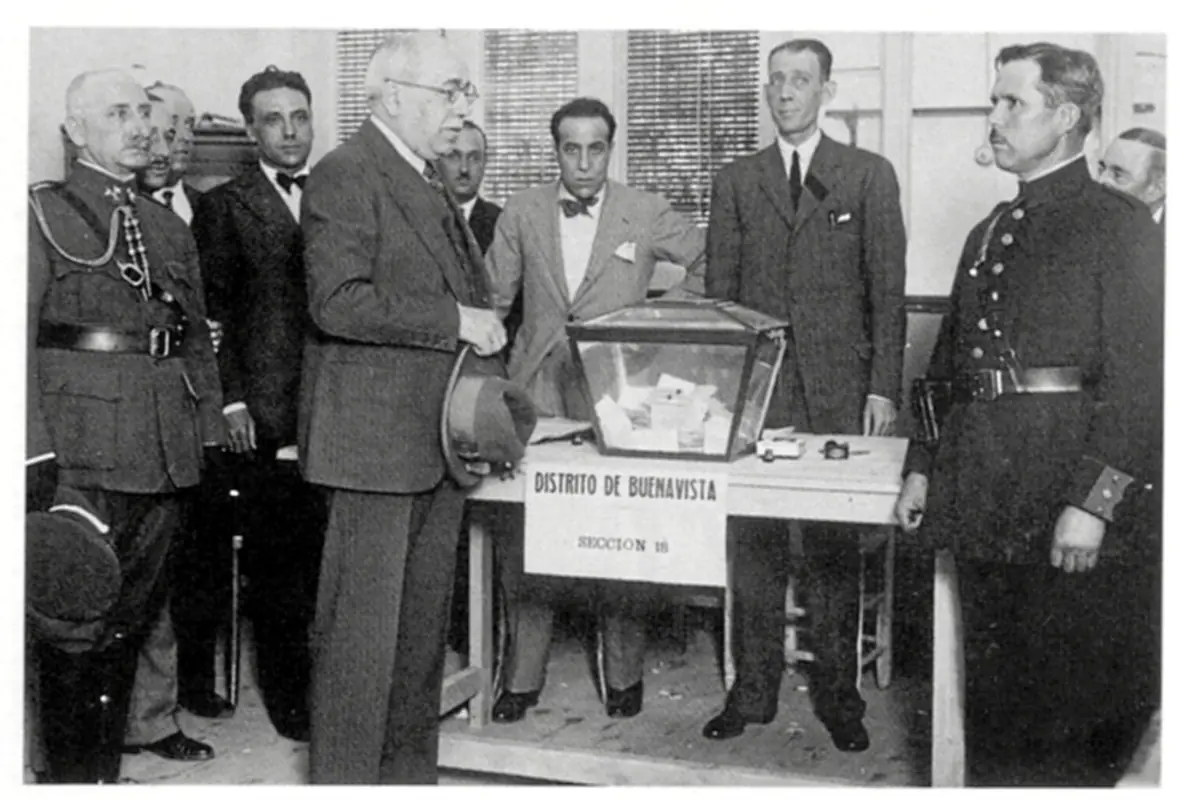 Fotografía histórica en blanco y negro de varios hombres con traje y uniformes militares reunidos alrededor de una urna de votación transparente en una mesa con el cartel Distrito de Buenavista