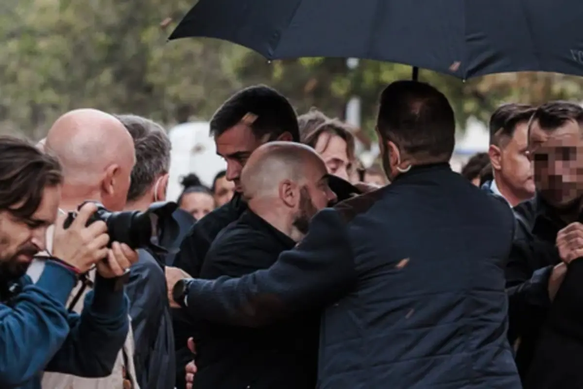 Grupo de personas en la calle bajo un paraguas oscuro mientras un fotógrafo captura la escena y varios hombres se empujan o sujetan entre sí