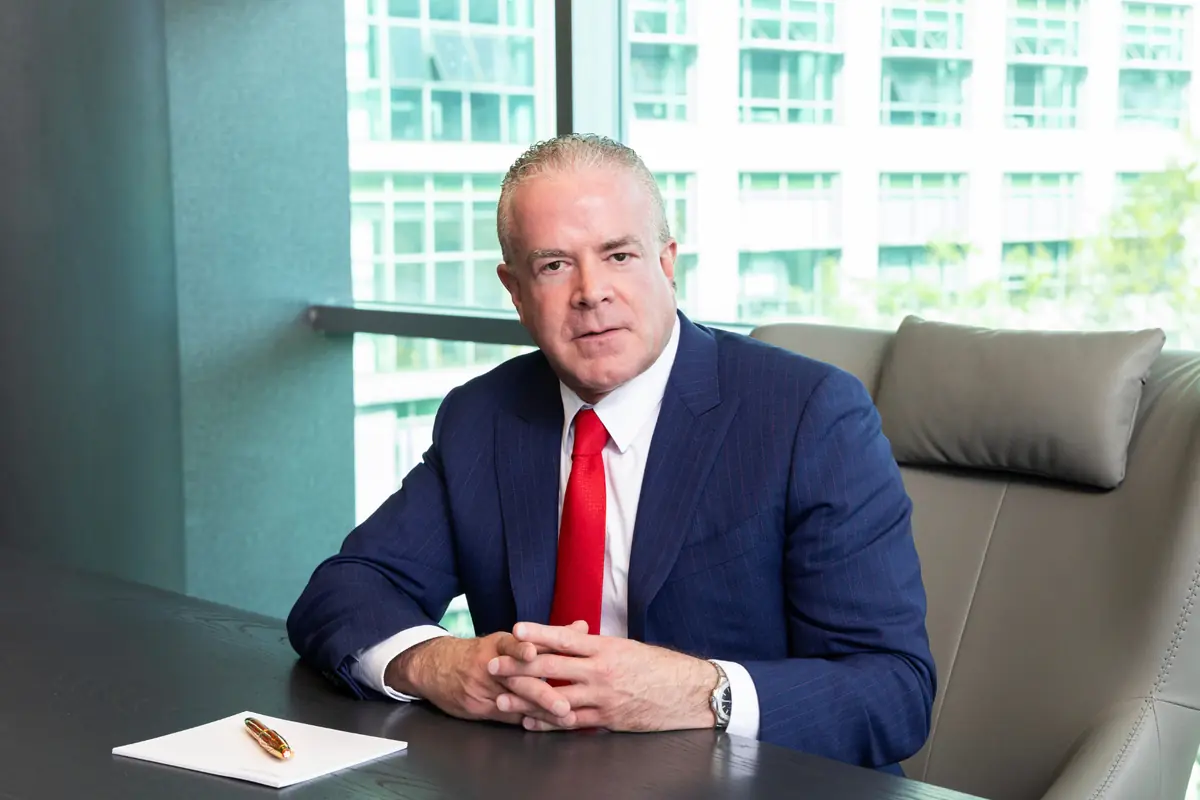 Hombre de mediana edad con traje azul y corbata roja sentado en una oficina moderna frente a una mesa con una libreta y una pluma