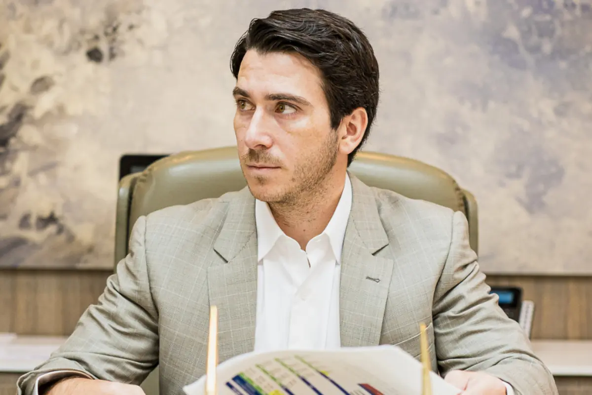 Hombre joven con traje claro sentado en una oficina mirando hacia un lado mientras sostiene documentos con gráficos de colores