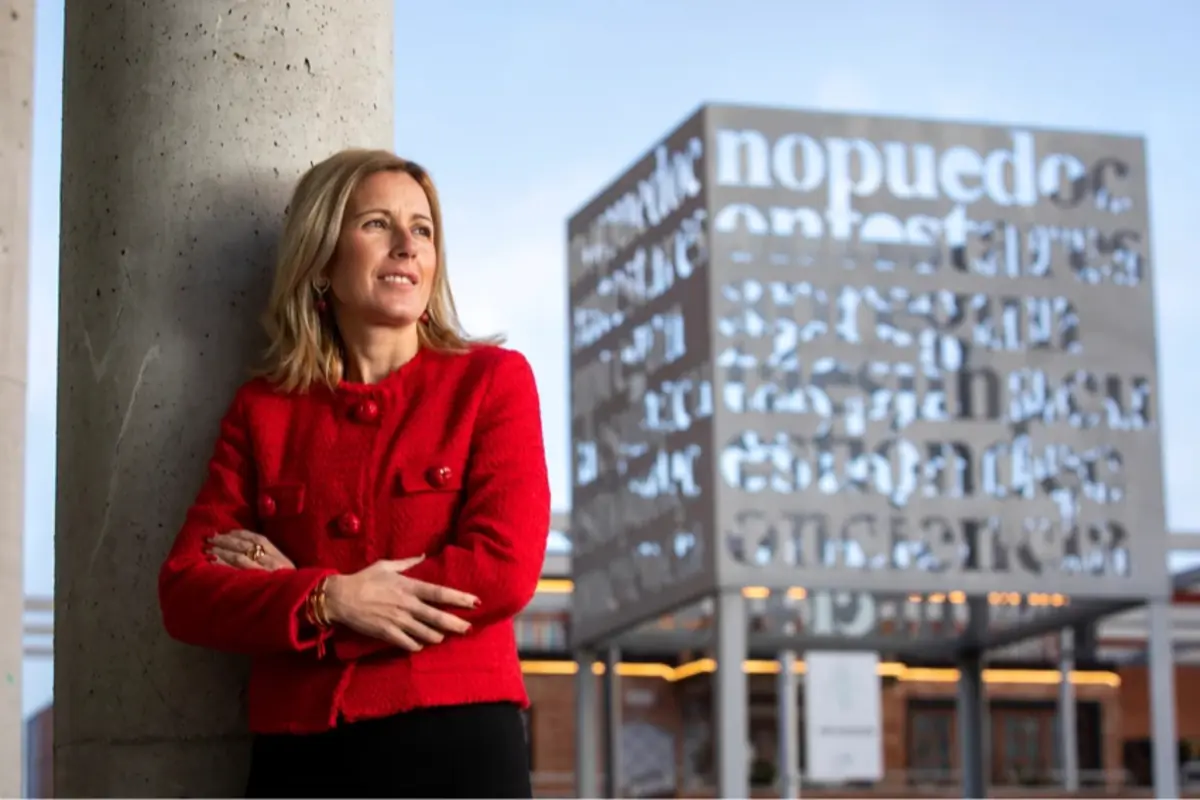 Mujer con chaqueta roja apoyada en una columna frente a una escultura cúbica metálica con letras recortadas en un espacio urbano