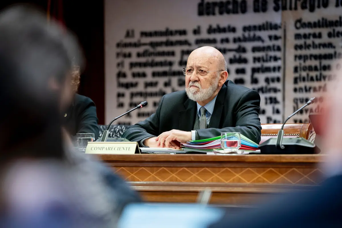 Hombre mayor con barba canosa sentado en una mesa de comparecencia parlamentaria con micrófonos y una carpeta de documentos de colores frente a él