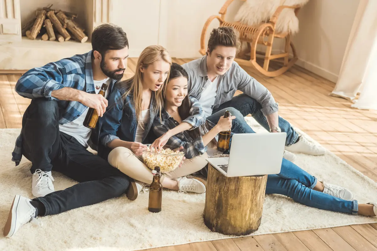 Grupo de amigos sentados en el suelo viendo una película en una laptop mientras comparten palomitas y bebidas en una sala acogedora