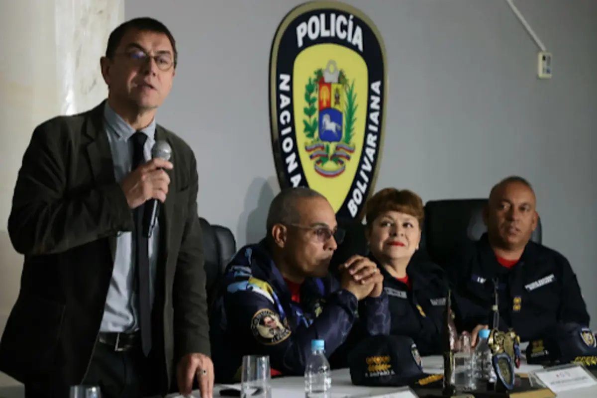 Cuatro autoridades policiales sentadas en una mesa y un hombre de traje hablando con micrófono frente a un escudo de la Policía Nacional Bolivariana Cuatro autoridades policiales sentadas en una mesa y un hombre de traje hablando con micrófono frente a un escudo de la Policía Nacional Bolivariana