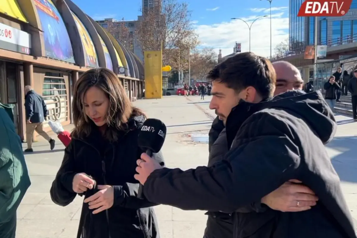 Reportero de EDA TV con micrófono entrevistando a una mujer en la calle mientras otra persona lo sujeta por detrás en una acera soleada de la ciudad