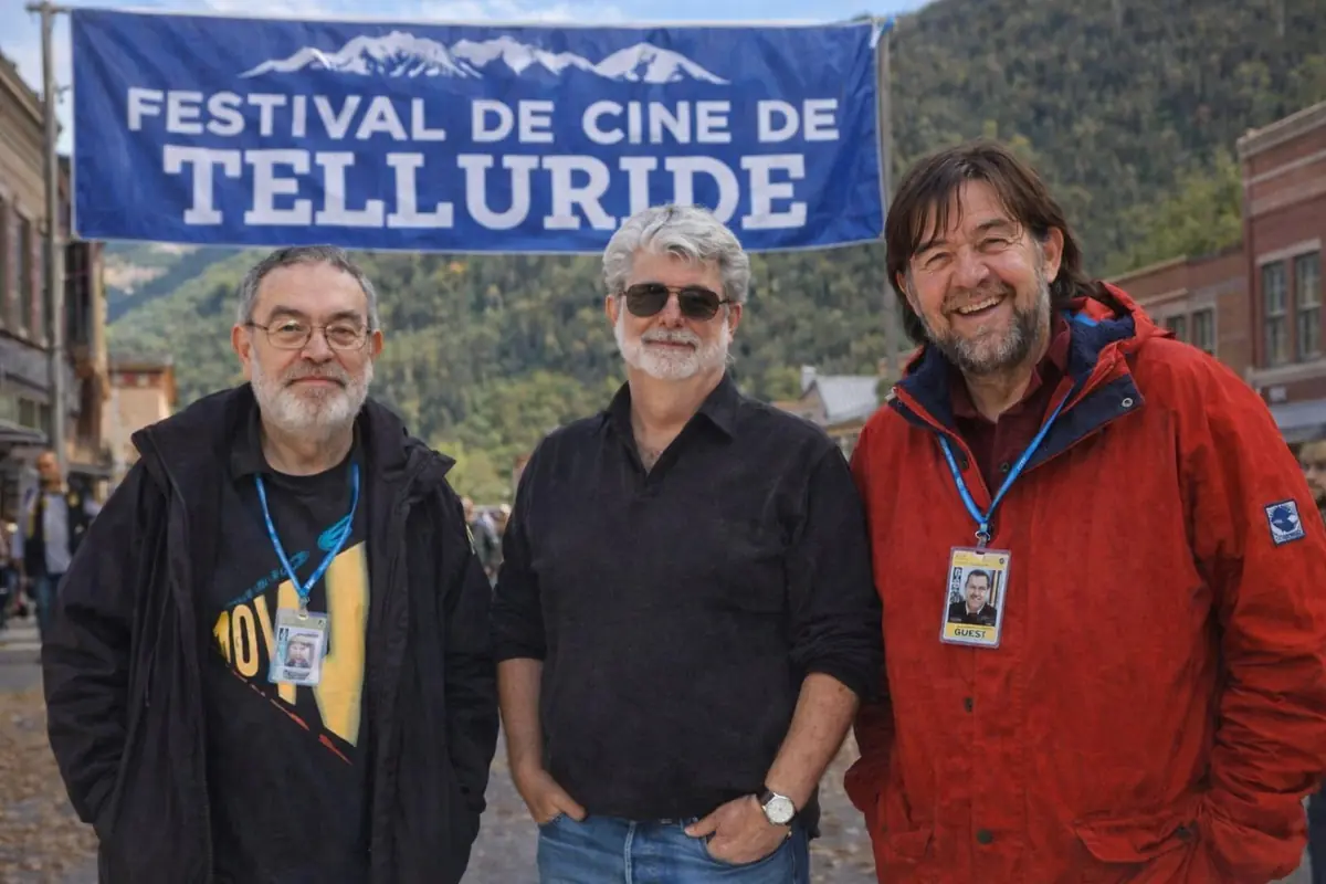 Tres hombres posan sonrientes frente a una calle al aire libre con un gran cartel azul del Festival de Cine de Telluride y montañas verdes al fondo