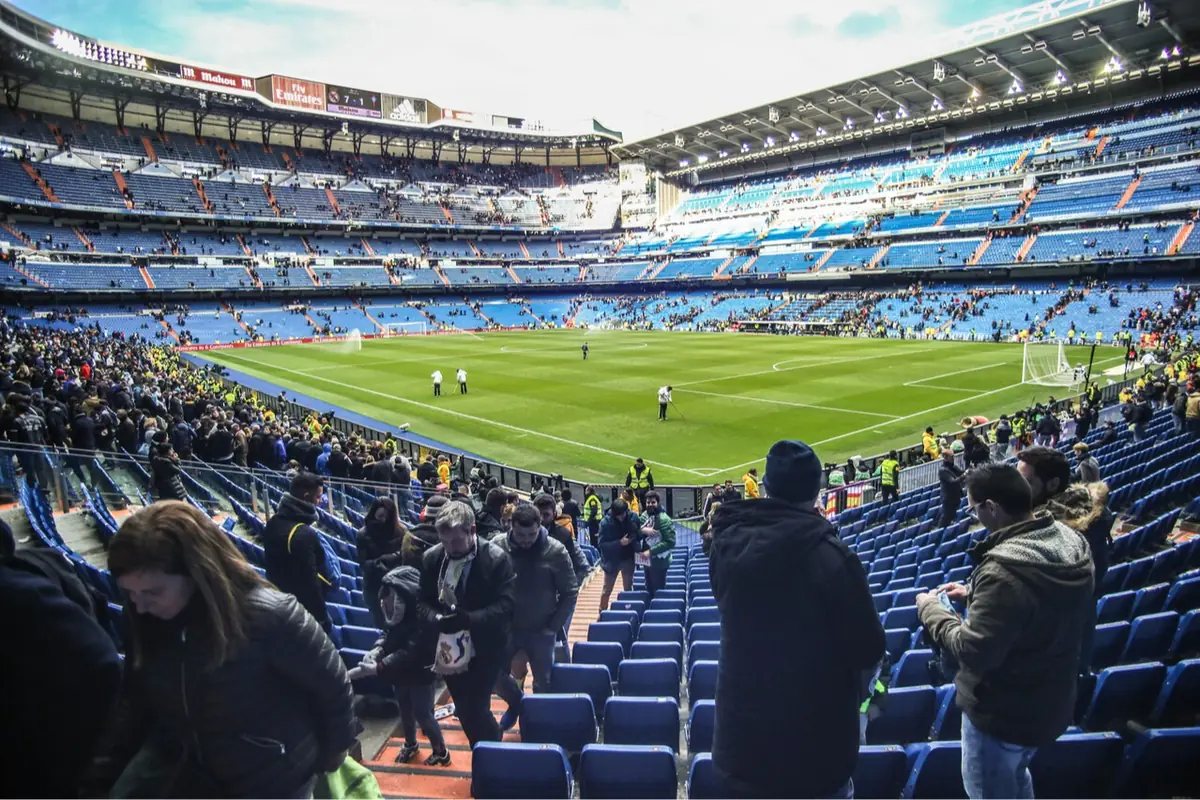 Aficionados entrando y tomando asiento en un gran estadio de fútbol con el campo verde listo para un partido