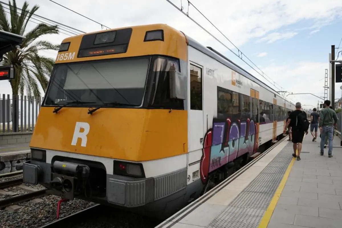 Tren de cercanías amarillo y blanco con grafitis en el lateral detenido en un andén mientras varias personas caminan por la estación