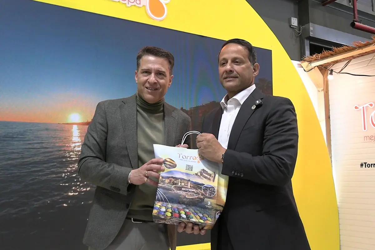 Dos hombres con traje posan sonrientes en un stand promocional sosteniendo una bolsa de regalo con imágenes turísticas frente a una pantalla con un atardecer sobre el mar