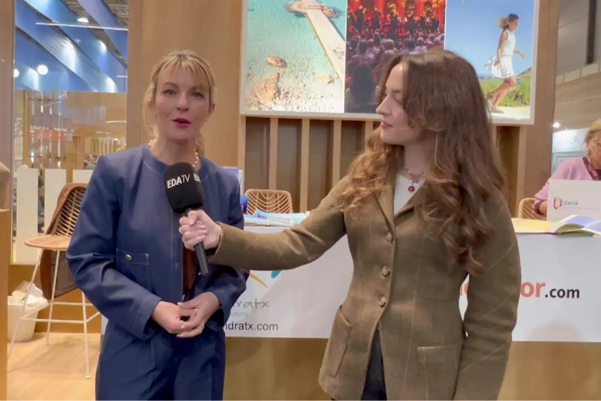 Reportera entrevistando a una mujer en un stand de feria turística mientras sostiene un micrófono de un canal de televisión