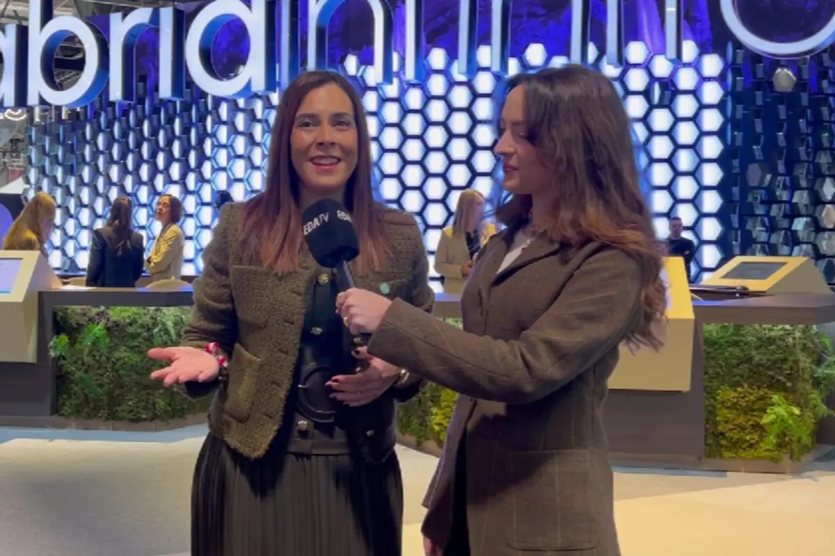 Dos mujeres en un stand de feria tecnológica mientras una de ellas sostiene un micrófono y entrevista a la otra frente a un fondo iluminado con paneles hexagonales