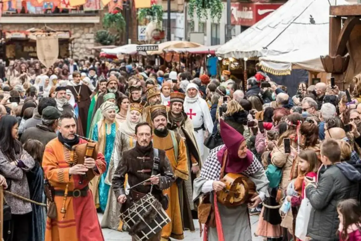 Desfile multitudinario en una feria medieval con músicos y personas disfrazadas de época avanzando entre puestos y público que observa y toma fotos