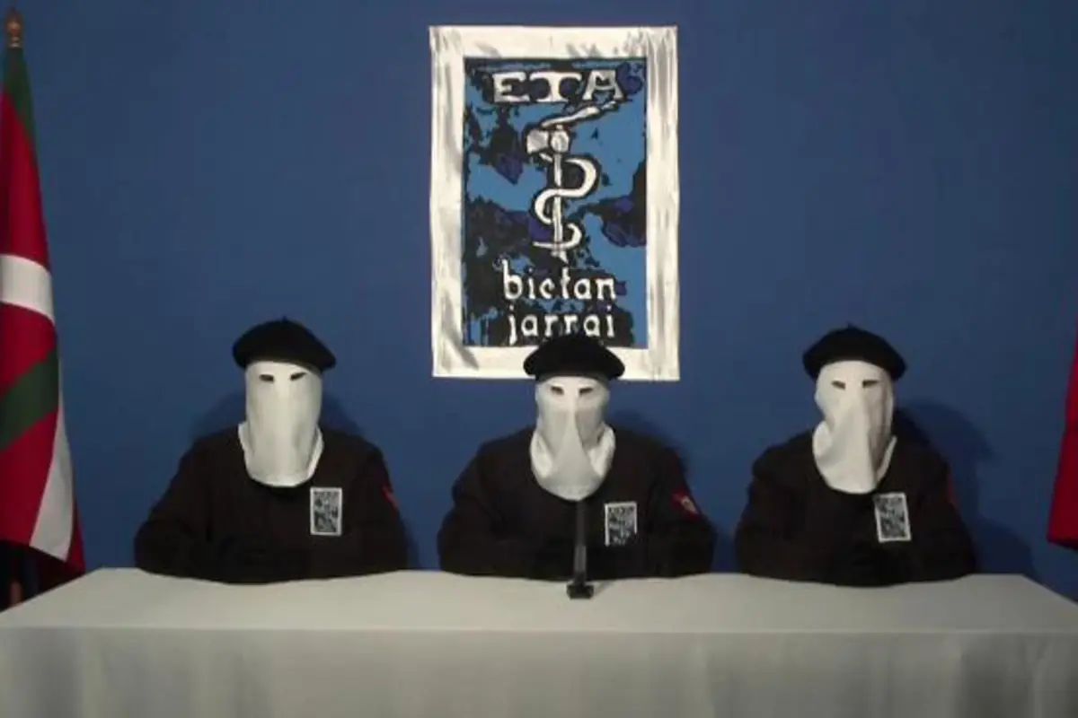 Tres personas enmascaradas con boinas negras sentadas tras una mesa frente a un fondo azul con un emblema en la pared