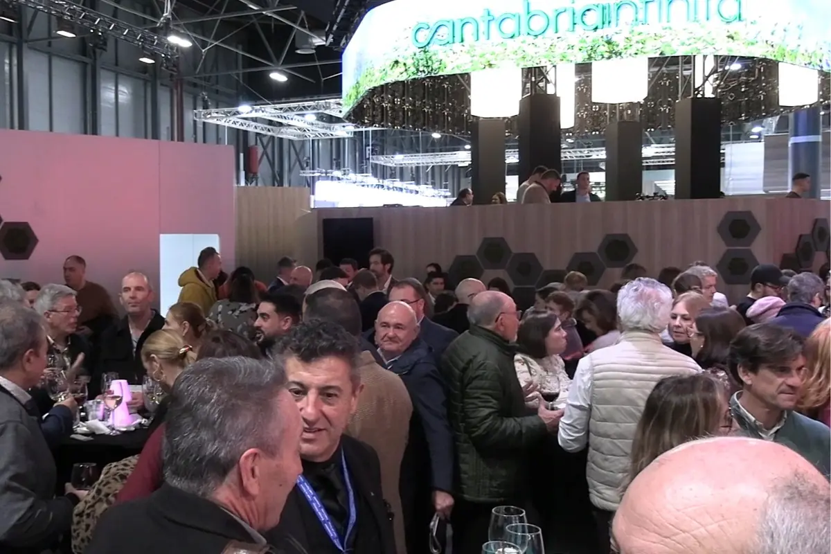 Multitud de personas conversando y tomando bebidas en un concurrido evento o feria en interiores con un gran stand iluminado de Cantabria al fondo