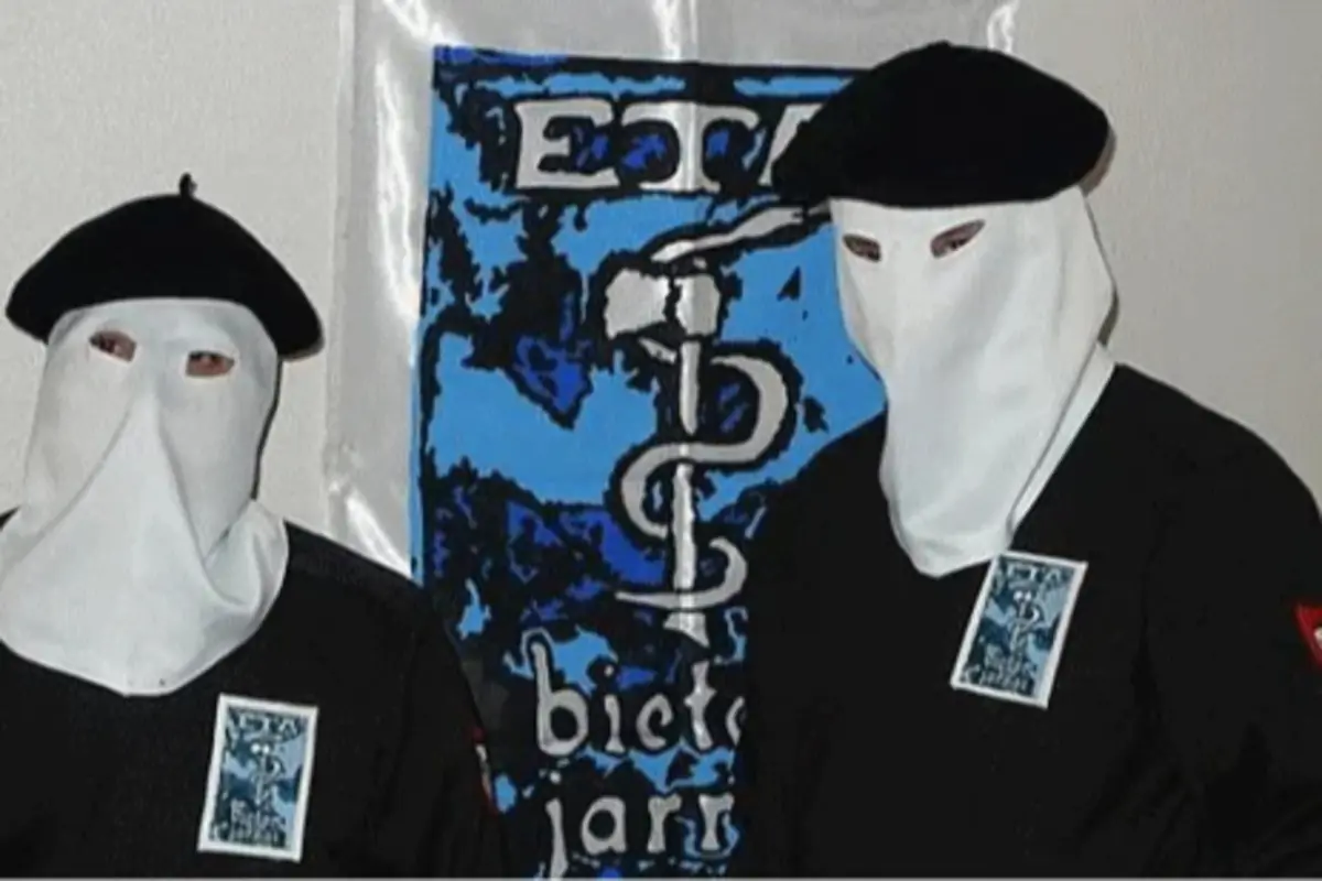 Dos personas encapuchadas con boinas negras posan frente a una gran pancarta azul con un emblema y letras en blanco