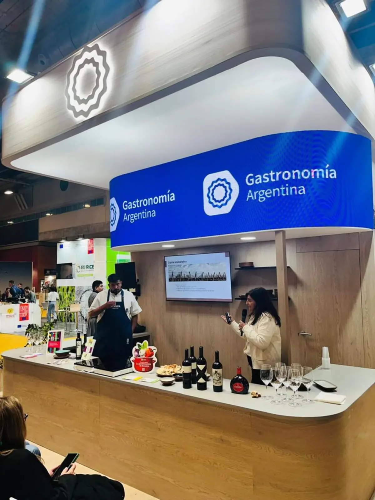 Stand de Gastronomía Argentina en una feria con una barra de madera donde se exhiben botellas de vino copas y aperitivos mientras una mujer presenta y un chef atiende al público