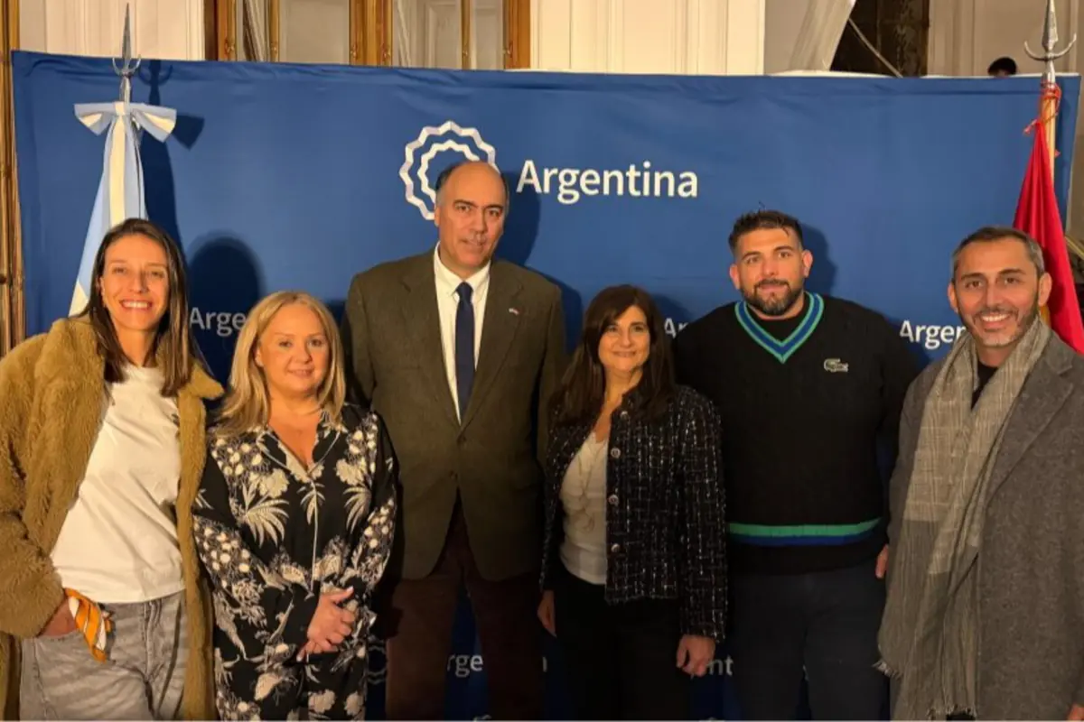 Seis personas posan sonrientes frente a un panel azul con la palabra Argentina y banderas a los costados en un entorno formal