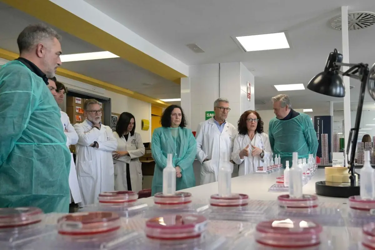 Grupo de personas con batas y equipos de protección observando una mesa llena de placas de laboratorio y frascos en un entorno científico