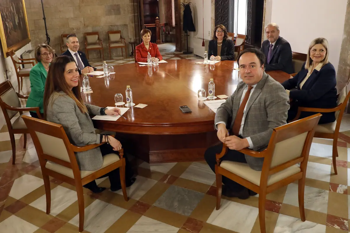 Varias personas sentadas alrededor de una gran mesa ovalada de madera en una sala de reuniones elegante con botellas de agua y documentos frente a ellas Varias personas sentadas alrededor de una gran mesa ovalada de madera en una sala de reuniones elegante con botellas de agua y documentos frente a ellas