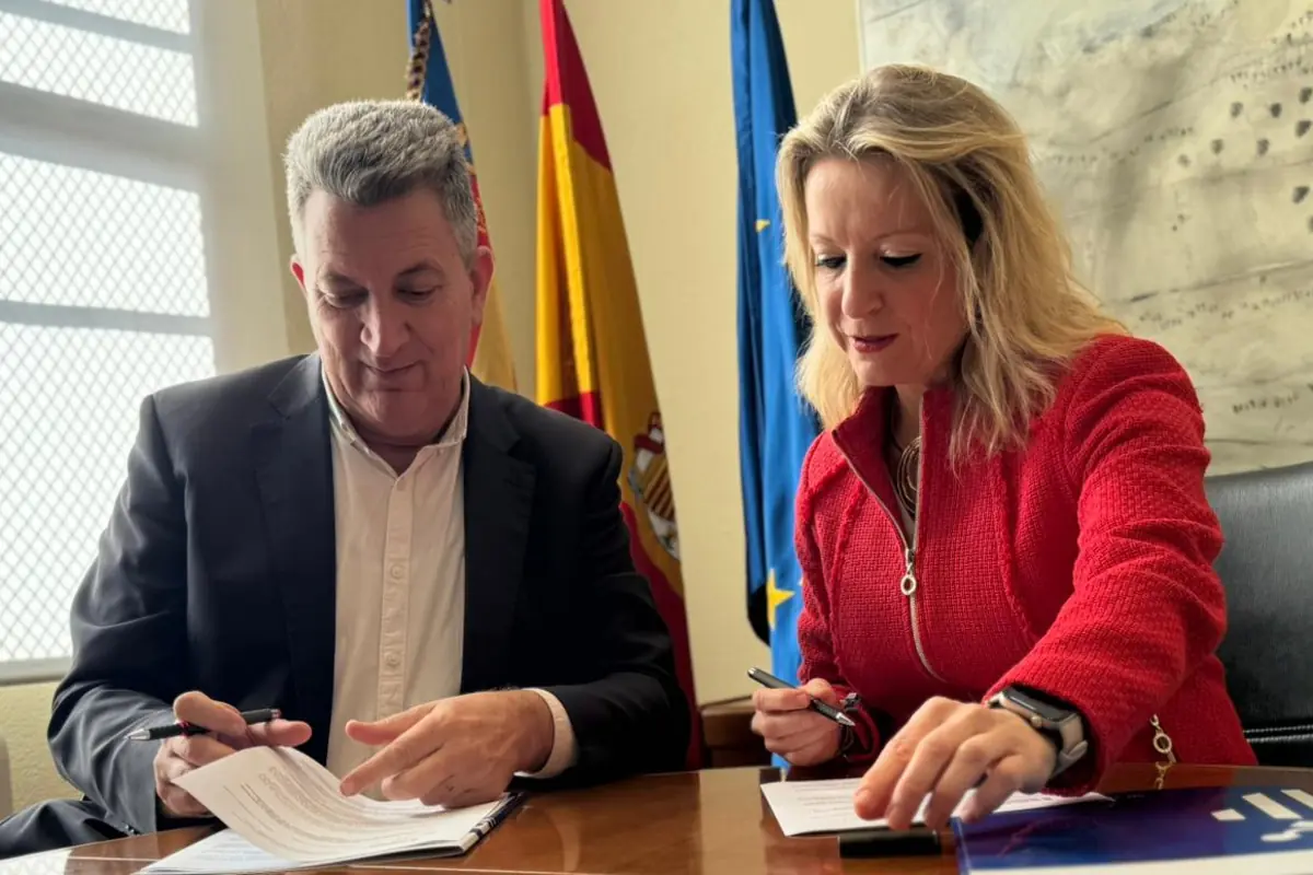 Dos personas sentadas en una mesa firmando documentos oficiales con banderas de España y de la Unión Europea al fondo
