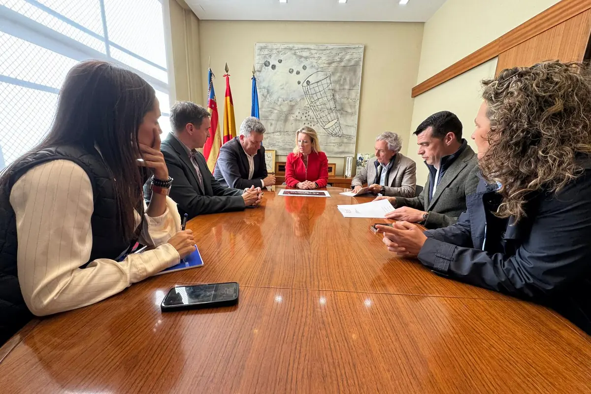 Reunión de seis personas sentadas alrededor de una mesa de madera en una oficina oficial con banderas al fondo mientras revisan documentos y toman notas