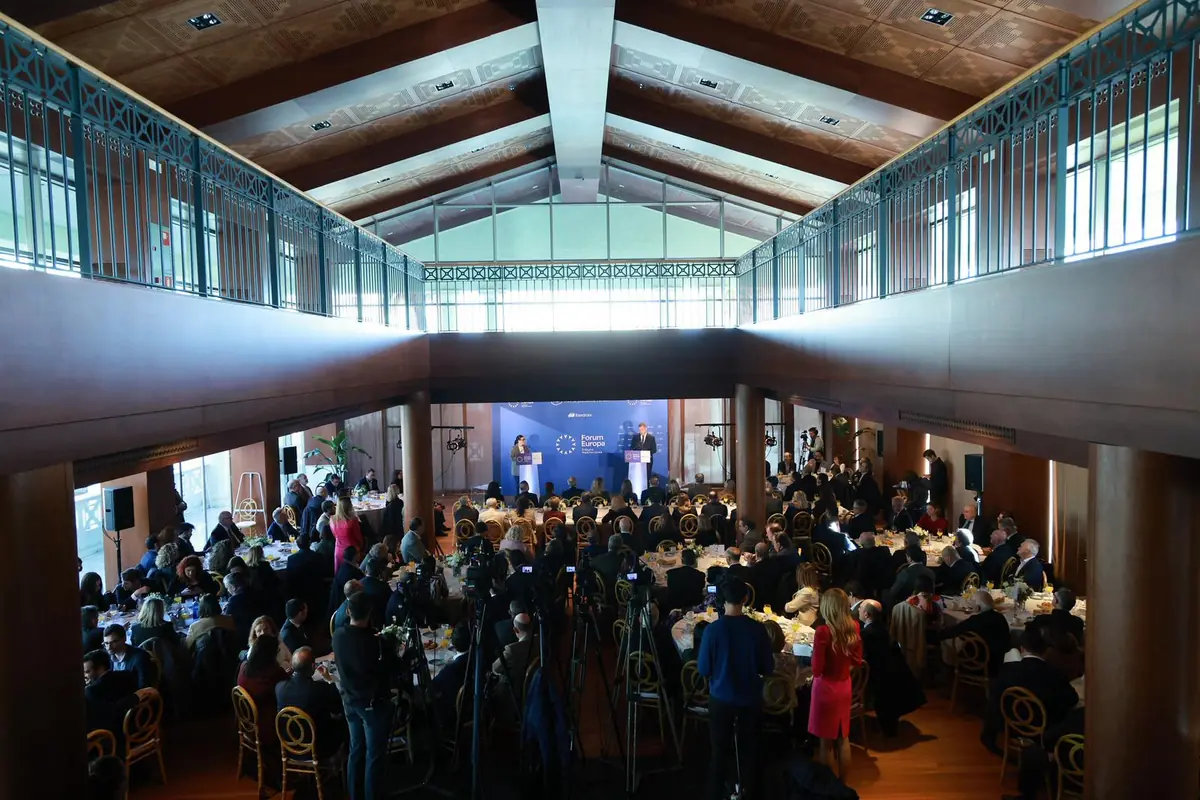 Gran salón con techo de madera lleno de mesas redondas y asistentes mientras dos ponentes hablan en un escenario al fondo durante un foro europeo Gran salón con techo de madera lleno de mesas redondas y asistentes mientras dos ponentes hablan en un escenario al fondo durante un foro europeo