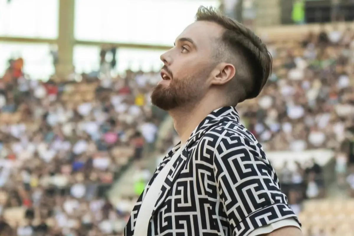 Hombre barbudo con camisa estampada en blanco y negro mirando hacia arriba en un estadio lleno de público desenfocado