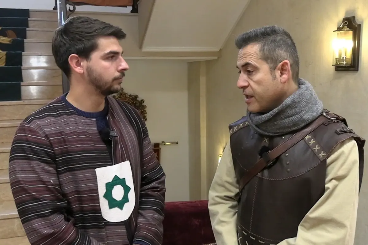 Dos hombres vestidos con trajes medievales conversan en el interior de un edificio junto a una escalera iluminada por apliques de pared Dos hombres vestidos con trajes medievales conversan en el interior de un edificio junto a una escalera iluminada por apliques de pared