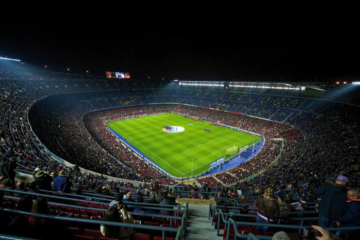 Estadio de fútbol iluminado de noche con las gradas llenas de aficionados y el campo preparado para un partido